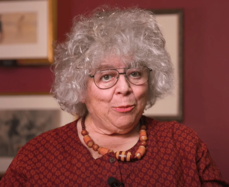 Miriam Margolyes in a red dress and wooden bead necklace, speaking to the camera